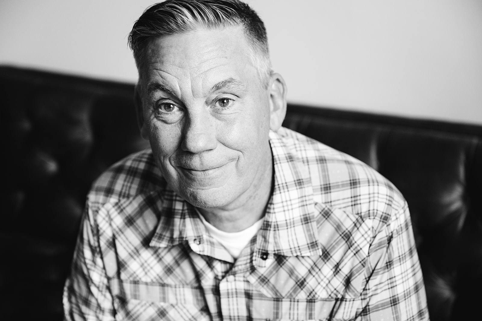 A man smiling while sitting on a leather couch, wearing a plaid shirt.